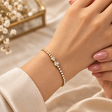 Double Pear Crystal Tennis Bracelet Gold Round Bezel - on wrist lifestyle shot: bracelet worn on elegant wrist against cream satin background with white florals, full round brilliant crystal tennis chain glowing with white brilliance, double pear crystal centre accent at front of wrist with exceptional three-dimensional sculptural presence