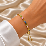 Gold Multi-Colour Emerald Cut Crystal Tennis Bracelet Bolo Adjustable Slider - hero flat lay on cream satin: full bracelet in circular form showing continuous warm 18K gold box-set multi-colour emerald-cut crystal tennis line cycling yellow citrine, sapphire blue, peridot green, and rose pink stones in open-sided gold cage settings, cream satin background