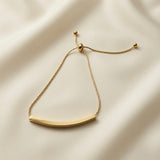 Lover Dream Curved Bar Bolo Bracelet - flat lay on ivory silk showing smooth mirror-polished gold curved bar, delicate cable chain, gold ball bolo slider and two gold ball tail ends