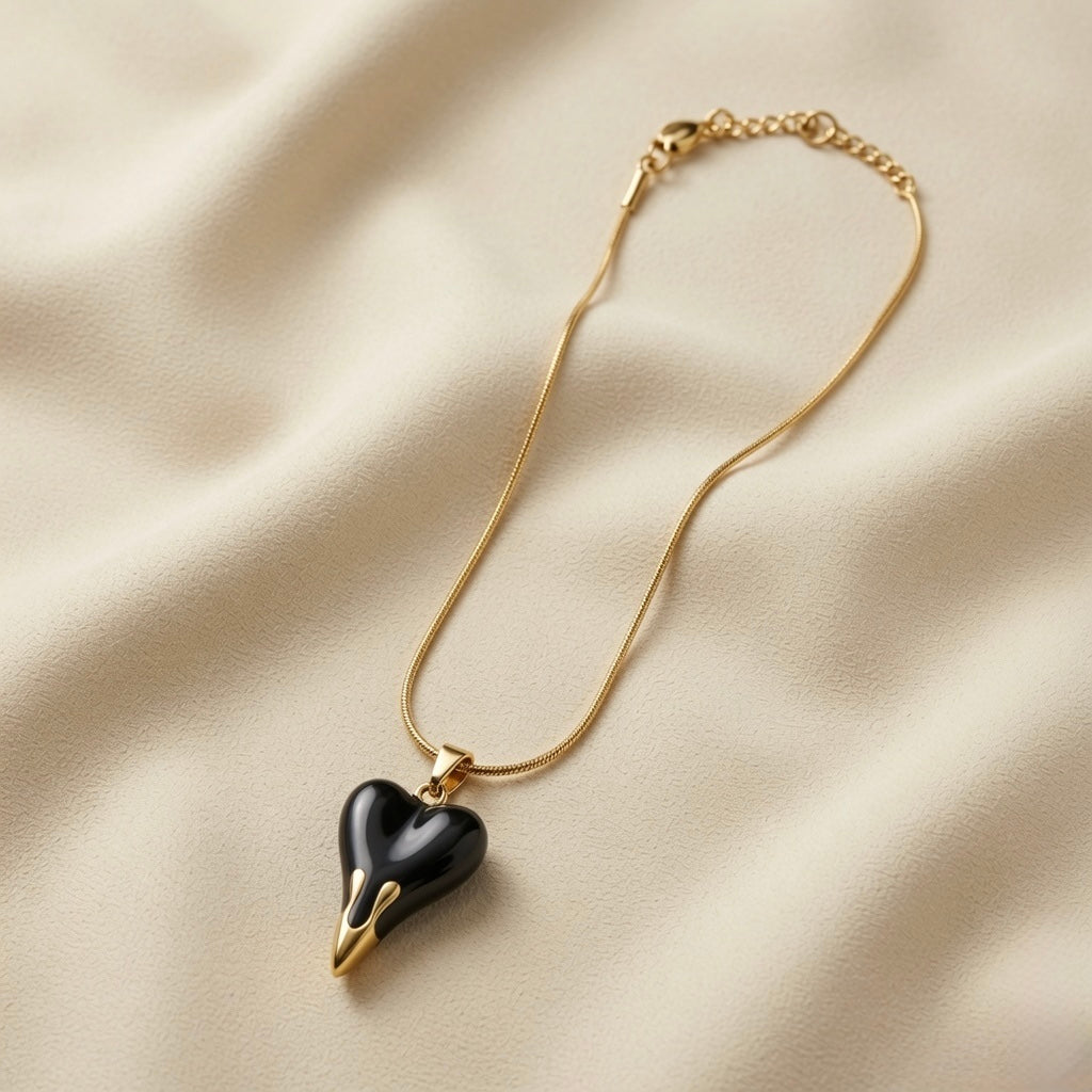 Coeur Noir Pendant Necklace - flat lay on ivory silk showing glossy black enamel 3D puffy heart with gold drip fang tips on flat snake chain