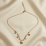 Cerise Charm Station Necklace - full flat lay on ivory silk showing complete snake chain with gold ball stations, five charms: cherry medallion, daisy, ruby pear, princess crystal, sapphire blue, and Svora logo bar tag on extender