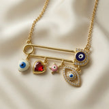 Amour Nazar Pin Charm Necklace - macro close-up on ivory silk showing large gold safety pin with navy pavé evil eye at clasp end and five sliding charms: white enamel evil eye heart, ruby red faceted crystal heart in gold bezel, pink enamel star, large pavé crystal marquise evil eye with blue centre