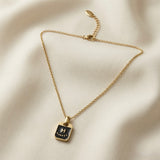 Initial Black Enamel Tag Pendant Necklace - full flat lay on ivory silk showing complete gold cable chain with lobster clasp, oval brand tag on curb extender and black enamel rectangular tag pendant with raised gold H initial