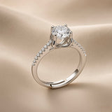 Lumière Solitaire Pavé Adjustable Ring - three-quarter view on champagne silk showing complete ring: round brilliant crystal zirconia solitaire in four-prong cathedral setting elevated above band, sculpted gallery beneath stone, pavé crystal zirconia accents along both shoulders on rhodium-plated silver-tone band with open-back adjustable mechanism at base