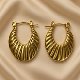 Soleil Fluted Oval Hoop Earrings - pair flat lay on champagne silk showing both wide oval hoop earrings with deep diagonal fluted ribbing across full face, hinged latch-back clasp with smooth gold pin and latch housing at top, warm 18K gold mirror-polish on raised ridges