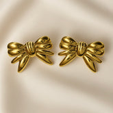 Ruban Puffy Bow Stud Earrings - pair front flat lay on ivory silk showing both large three-dimensional puffy bow stud earrings in warm 18K gold: two rounded lobes at top, tightly gathered ribbed knot at centre with three horizontal grooves, four splayed ribbon tail strands fanning outward and downward