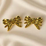 Ruban Puffy Bow Stud Earrings - pair front flat lay on ivory silk showing both large three-dimensional puffy bow stud earrings in warm 18K gold from slightly different angle: rounded lobes, ribbed central knot and four splayed ribbon tails with deep shadow recesses between elements, mirror-polish warm gold surface