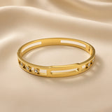 Arcade Crystal Lattice Hinged Bangle Set - crystal bangle three-quarter overhead view on champagne silk showing wide flat-band hinged bangle in warm 18K gold with open-work geometric lattice of alternating rectangular and oval cut-outs, five bezel-set round crystal zirconia stones in smooth cylindrical gold bezels evenly distributed across front arc, hinged clasp at back