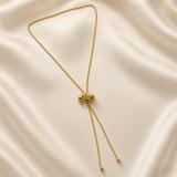 Grand Ruban Bow Lariat Box Chain Necklace - full flat lay on ivory silk showing complete necklace: continuous square-link gold box chain loop with large three-dimensional puffy bow slider at centre, two tail chains descending with smooth polished gold ball finials at ends, bow showing two rounded lobes, ribbed knot and four splayed ribbon tail strands