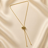 Grand Ruban Bow Lariat Box Chain Necklace - full flat lay on ivory silk showing complete necklace from slightly different angle: continuous square-link gold box chain loop with large puffy bow slider at centre showing two rounded lobes and ribbed knot, two parallel tail chains with smooth polished gold ball finials descending below bow