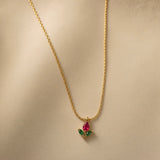 Tulipe Rouge Crystal Bud Pendant Necklace - three-quarter close-up on warm beige background showing red tulip bud pendant in full detail: pear-cut deep ruby red crystal bud in minimal gold bezel set point-downward, two marquise-cut deep forest green crystal leaves set horizontally outward from fine gold stem, smooth gold bail at top, fine twisted gold cable chain above