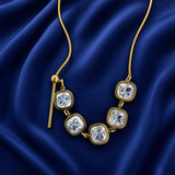 Lumière Cinq Cushion Crystal Convertible Snake Chain Necklace - three-quarter close-up on deep navy velvet showing five cushion-cut crystal zirconia station arc: five generous cushion-cut diamond-clear crystals in smooth gold bezels linked in continuous arc, gold bar-and-ball toggle at left, flat-link snake chain on both sides, pure white diamond-like brilliance of cushion crystals clearly visible