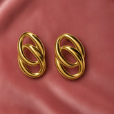 Gold Double Oval Interlocked Link Stud Earrings - pair face-on on dusty pink satin showing complete earrings from slightly wider angle: vertical oval silhouette with large outer oval and smaller inner oval interlocked, high mirror-polish warm gold surface reflecting dusty pink satin background, bold statement scale and three-dimensional chain-link construction clearly visible
