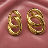 Gold Double Oval Interlocked Link Stud Earrings - pair elevated three-quarter close-up on dusty pink satin showing full three-dimensional depth of double oval interlock: large outer oval tube and smaller inner oval tube passing through each other, thick rounded tube walls in high mirror-polish gold, warm gold highlights and shadows of interlocked oval composition, straight post with butterfly clutch back visible on right earring