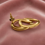 Gold Double Oval Interlocked Link Stud Earrings - side view on dusty pink satin showing single earring three-dimensional construction: large outer oval tube and smaller inner oval tube genuinely interlocked in full 3D, thick rounded tube walls in mirror-polish gold, straight gold post with butterfly friction clutch back visible at top rear