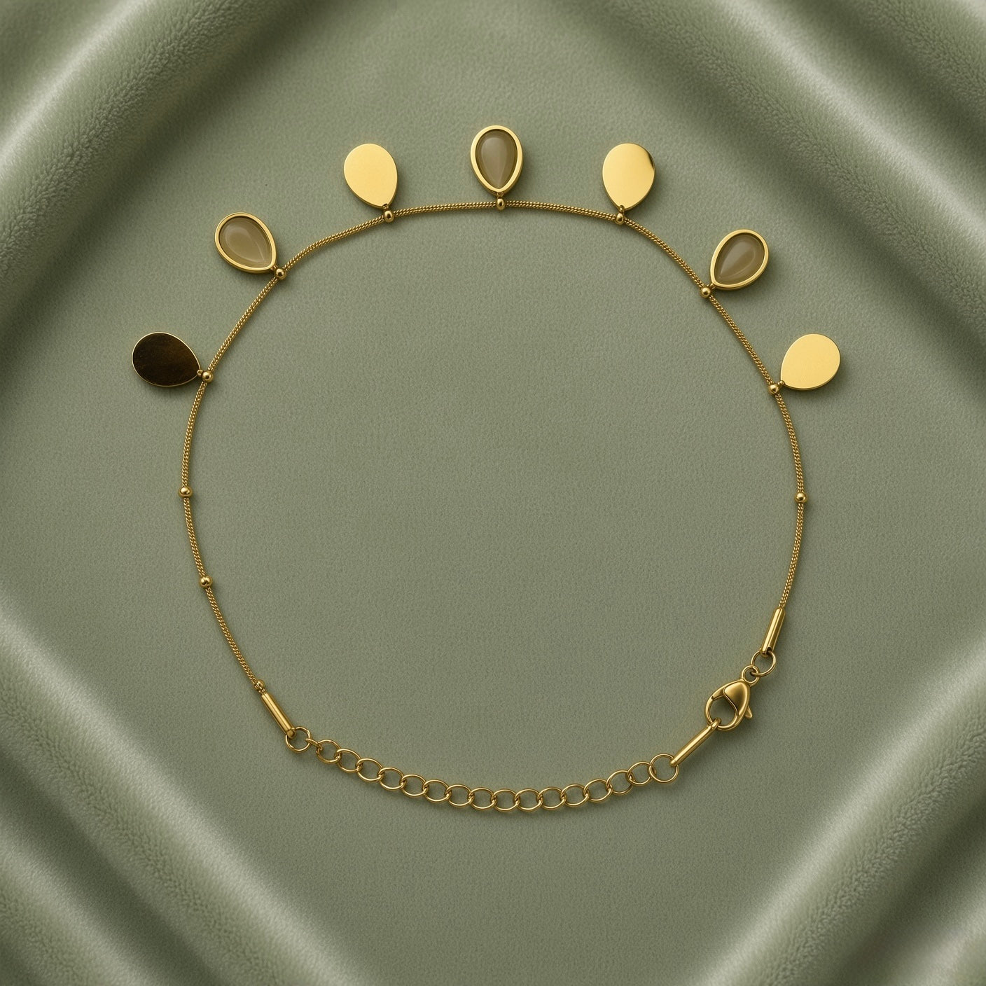Gold Teardrop Charm Satellite Chain Anklet - full flat lay on sage green satin tray showing complete anklet in circular layout: fine gold satellite chain with evenly spaced gold ball stations carrying seven alternating teardrop charms along upper arc, solid mirror-polish gold flat teardrop charms alternating with open gold bezel nude cabochon teardrop charms, lobster clasp and curb-link extender chain at bottom right