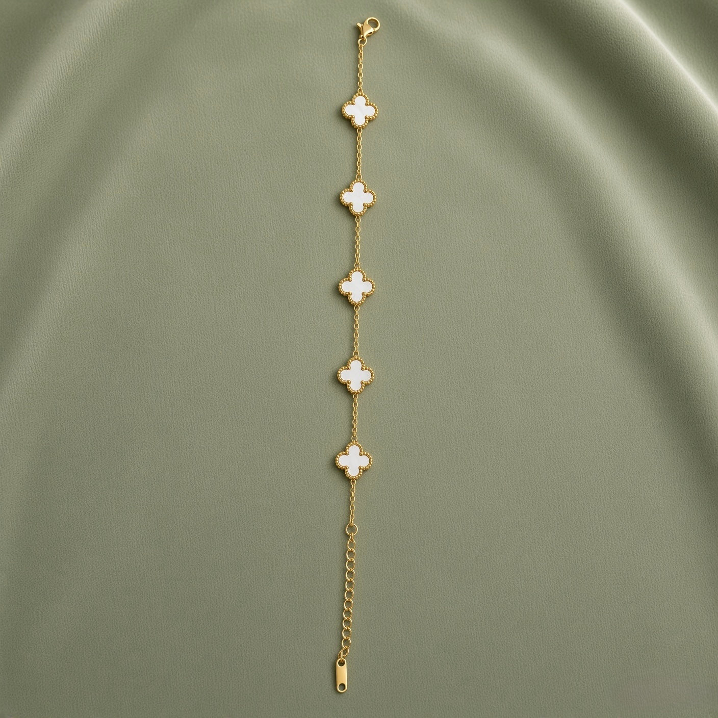 White Clover Station Bracelet Gold Milgrain - full length flat lay on sage green satin showing complete bracelet laid straight: five white mother-of-pearl four-leaf clover motifs with continuous milgrain gold bead borders evenly spaced on fine gold cable link chain, lobster clasp at top, curb-link extender chain and gold barrel tag at bottom
