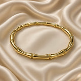 Gold Bamboo Bangle Bracelet Mirror Polish Sculptural - overhead flat lay on cream satin: top-down view of full circular bangle showing all bamboo stalk segments and double-ring joint nodes in mirror-polish warm gold, even warm gold tone across all tube sections, cream satin background