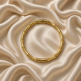 Gold Bamboo Bangle Bracelet Mirror Polish Sculptural - angled flat lay on cream satin: three-quarter angled view of bamboo gold bangle showing segment depth and node joint detail, mirror-polish warm 18K gold surface catching directional light across bamboo tube crown highlights, cream satin background