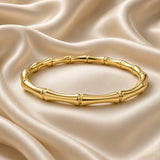 Gold Bamboo Bangle Bracelet Mirror Polish Sculptural - on wrist lifestyle: bangle worn on elegant wrist with white sleeve, bamboo motif tube sections and double-ring node joints visible in warm mirror-polish gold wrapping wrist, hinge gap visible at top, slim elegant wrist profile, white satin background