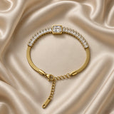 Gold Herringbone Chain Bracelet Baguette Crystal Station Emerald Bezel Centre - overhead circular flat lay on cream satin: full bracelet in circular form showing complete herringbone chain body, full baguette crystal channel row, large emerald bezel centre at top, lobster clasp and adjustable curb link extension chain at bottom, cream satin background