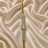 Gold Herringbone Chain Bracelet Baguette Crystal Station Emerald Bezel Centre - vertical detail flat lay on cream satin: straight layout showing full baguette crystal station row with large emerald bezel centre, individual channel-set rectangular baguette stones visible in continuous row, large emerald-cut centre in rounded-corner warm gold bezel, flat herringbone chain at top, lobster clasp and extension chain at bottom, cream satin background