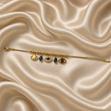 Gold Evil Eye Charm Bracelet Blue Crystal Teardrop Pavé Nazar Snake Chain - angled flat lay on cream satin: three-quarter view of bracelet showing charm section detail with alternating bead stations and all 5 suspended charms at angle showing charm depth and individual charm construction, textured rib barrel beads and smooth gold round beads on snake chain, cream satin background