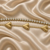 Gold Double Strand Bracelet Crystal Tennis Row Triple Puffy Heart Charm Chain - macro detail flat lay on cream satin: extreme close-up showing upper round brilliant white crystal zirconia tennis row with individually cup-set stones edge-to-edge and lower strand with three volumetric mirror-smooth domed warm gold puffy heart charms with heart cleft at top, fine warm gold cable chain between hearts, warm gold sculptural detail of puffy heart surfaces visible, cream satin background