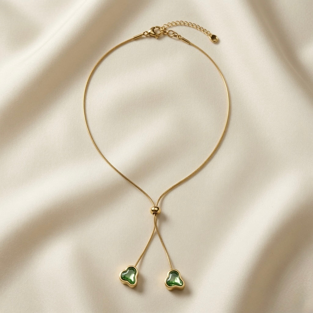 Émeraude Coeur Lariat Necklace - full flat lay on ivory silk showing snake chain, gold bead slider and two emerald heart pendants
