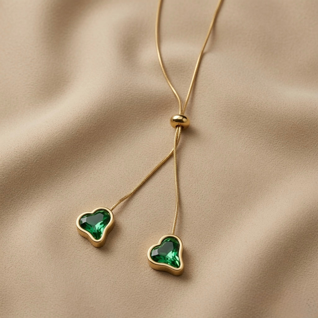 Émeraude Coeur Lariat Necklace - macro on sand showing asymmetric emerald green crystal hearts in gold bezels at different drop lengths
