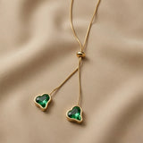 Émeraude Coeur Lariat Necklace - macro on sand showing asymmetric emerald green crystal hearts in gold bezels at different drop lengths
