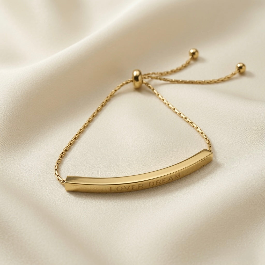 Lover Dream Curved Bar Bolo Bracelet - close-up flat lay on ivory silk showing LOVER DREAM engraving on curved gold bar face, bolo slider mechanism and cable chain detail