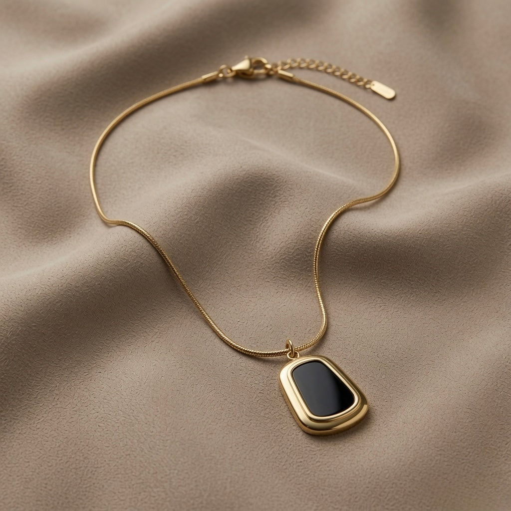 Tableau Noir Pendant Necklace - flat lay on taupe silk showing black enamel rounded-rectangle pendant in wide gold stepped frame on snake chain