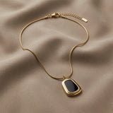 Tableau Noir Pendant Necklace - flat lay on taupe silk showing black enamel rounded-rectangle pendant in wide gold stepped frame on snake chain