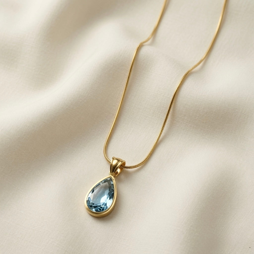 Larme Crystal Pear Pendant Necklace Aquamarine - flat lay on ivory silk showing faceted pear-cut aquamarine crystal in gold bezel on snake chain
