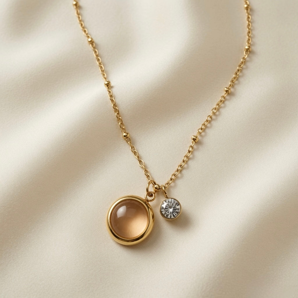 Lune Dorée Crystal Duo Pendant Necklace - close-up flat lay on champagne silk showing warm champagne-peach domed cabochon in deep gold bezel and small brilliant crystal side by side at slightly different heights