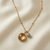 Lune Dorée Crystal Duo Pendant Necklace - close-up flat lay on champagne silk showing warm champagne-peach domed cabochon in deep gold bezel and small brilliant crystal side by side at slightly different heights