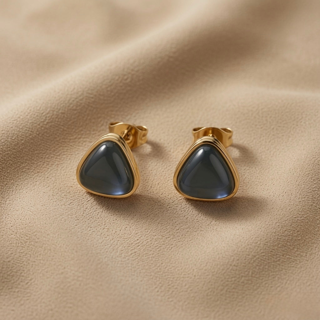 Orage Bleu Triangle Earrings - macro close-up on taupe silk showing pair of rounded-triangle slate blue-grey cabochon studs in ridged gold bezel with butterfly push-backs