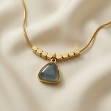 Orage Bleu Triangle Necklace - macro close-up on ivory silk showing faceted gold hexagonal beads, ridged gold bezel and slate blue-grey triangle cabochon pendant detail