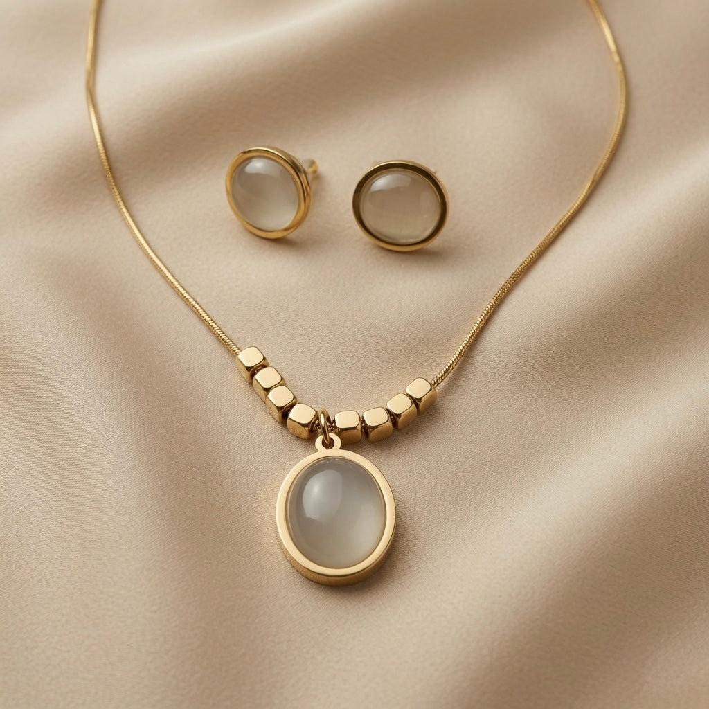 Lune Blanche Cabochon Jewellery Set - flat lay on champagne silk showing matching oval pendant necklace with gold cube beads and round stud earrings in white moonstone-effect cabochon