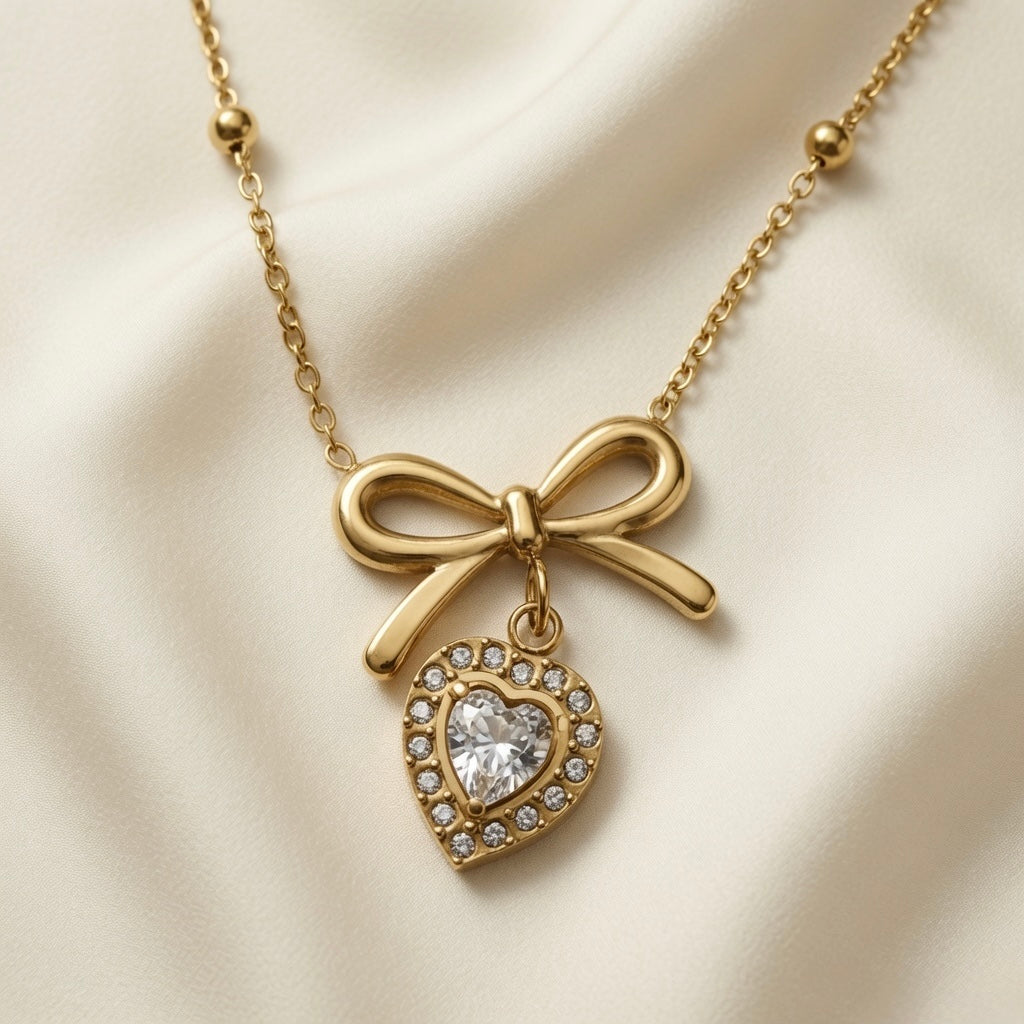 Noeud Coeur Crystal Bow Necklace - close-up flat lay on ivory silk showing sculptural gold bow pendant with dangling heart-cut crystal zirconia pavé halo heart on satellite chain