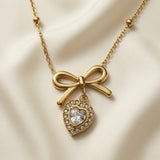 Noeud Coeur Crystal Bow Necklace - close-up flat lay on ivory silk showing sculptural gold bow pendant with dangling heart-cut crystal zirconia pavé halo heart on satellite chain
