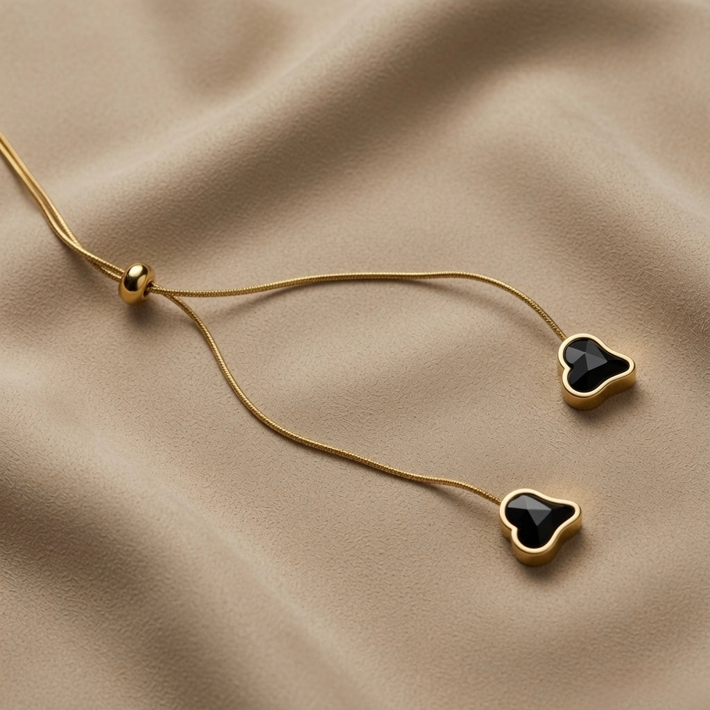Coeur Noir Bolo Lariat Necklace - close-up on taupe silk showing gold ball slider mechanism, two snake chain tails descending and two black crystal heart pendants at different lengths