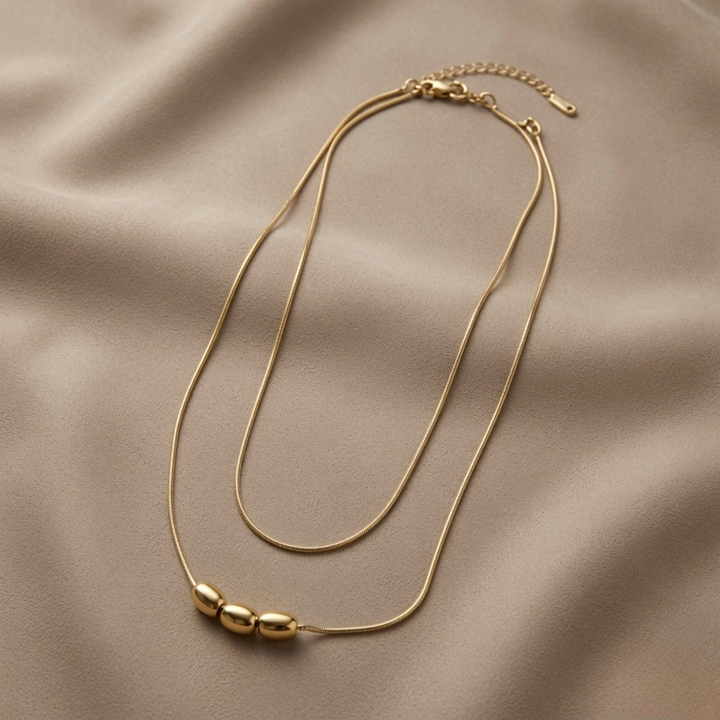 Trio Ovale Double Layer Snake Chain Necklace - flat lay on taupe silk showing double snake chain with three mirror-polished gold oval barrel beads on lower chain