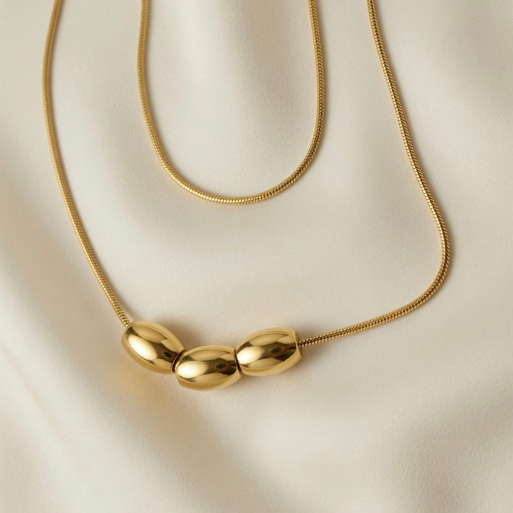 Trio Ovale Double Layer Snake Chain Necklace - macro close-up on ivory silk showing three mirror-polished gold oval barrel beads reflecting light on lower snake chain