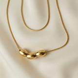 Trio Ovale Double Layer Snake Chain Necklace - macro close-up on ivory silk showing three mirror-polished gold oval barrel beads reflecting light on lower snake chain