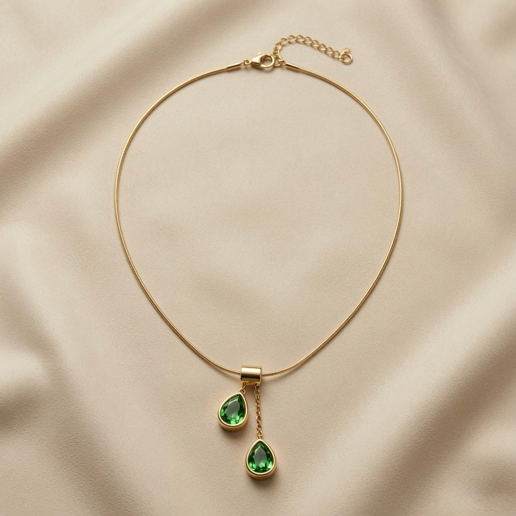 Duo Larme Émeraude Asymmetric Pendant Necklace - full flat lay on ivory silk showing rectangular gold bail, two asymmetric emerald green pear crystal pendants and flat snake chain with lobster clasp extender