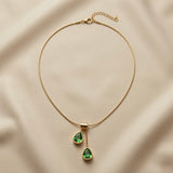 Duo Larme Émeraude Asymmetric Pendant Necklace - full flat lay on ivory silk showing rectangular gold bail, two asymmetric emerald green pear crystal pendants and flat snake chain with lobster clasp extender
