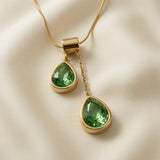 Duo Larme Émeraude Asymmetric Pendant Necklace - macro close-up on ivory silk showing two bezel-set emerald green pear-cut crystals at asymmetric heights, gold bezels and cable chain drop detail
