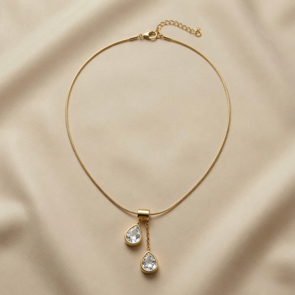 Duo Larme Crystal Asymmetric Pendant Necklace - full flat lay on champagne silk showing complete snake chain with lobster clasp extender, rectangular gold bail and two asymmetric white crystal pear pendants